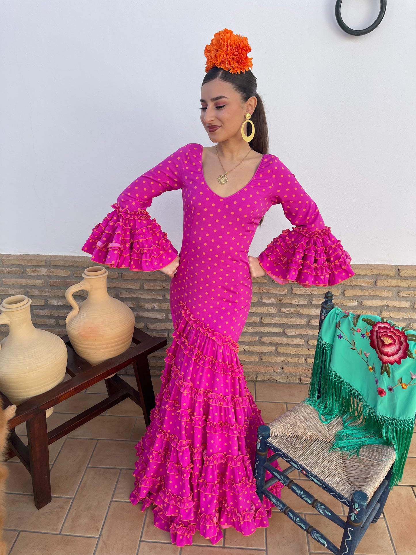 Traje de flamenca mujer 2024 rosa fucsia con lunares naranja y de corte canastero. De complementos se pueden ver en la cabeza clavel naranja, pendientes de flamenca dorado y mantón verde agua con flores bordadas.