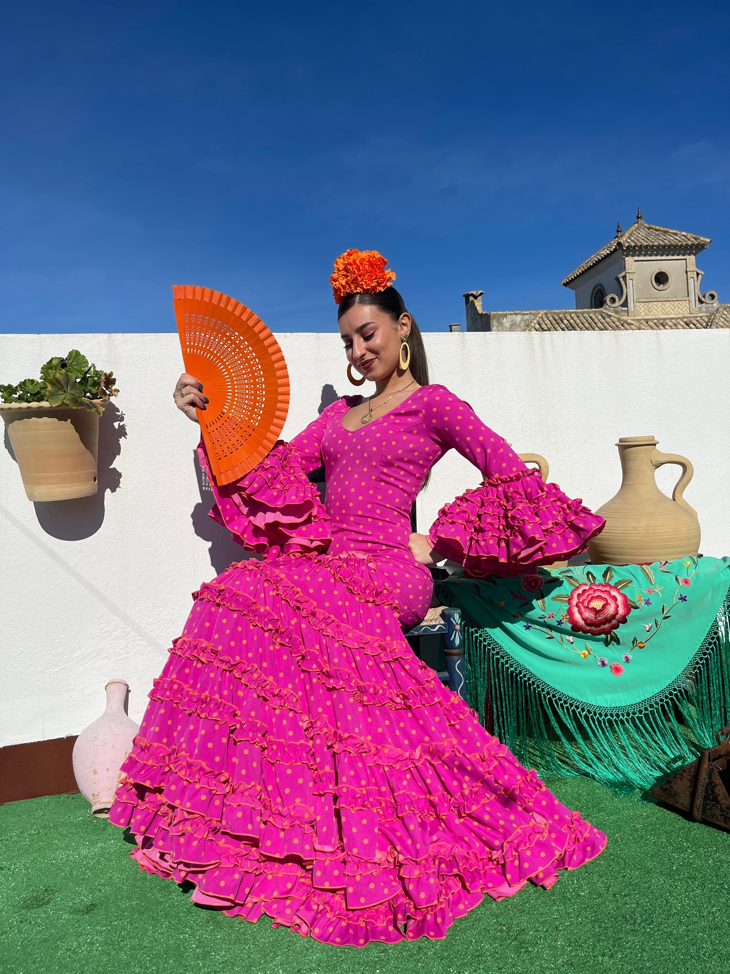 Traje de flamenca mujer 2024 rosa fucsia con lunares naranja y de corte canastero. De complementos se pueden ver un abanico naranja, claveles naranjas, pendientes de flamenca dorado y mantón verde agua con flores bordadas.