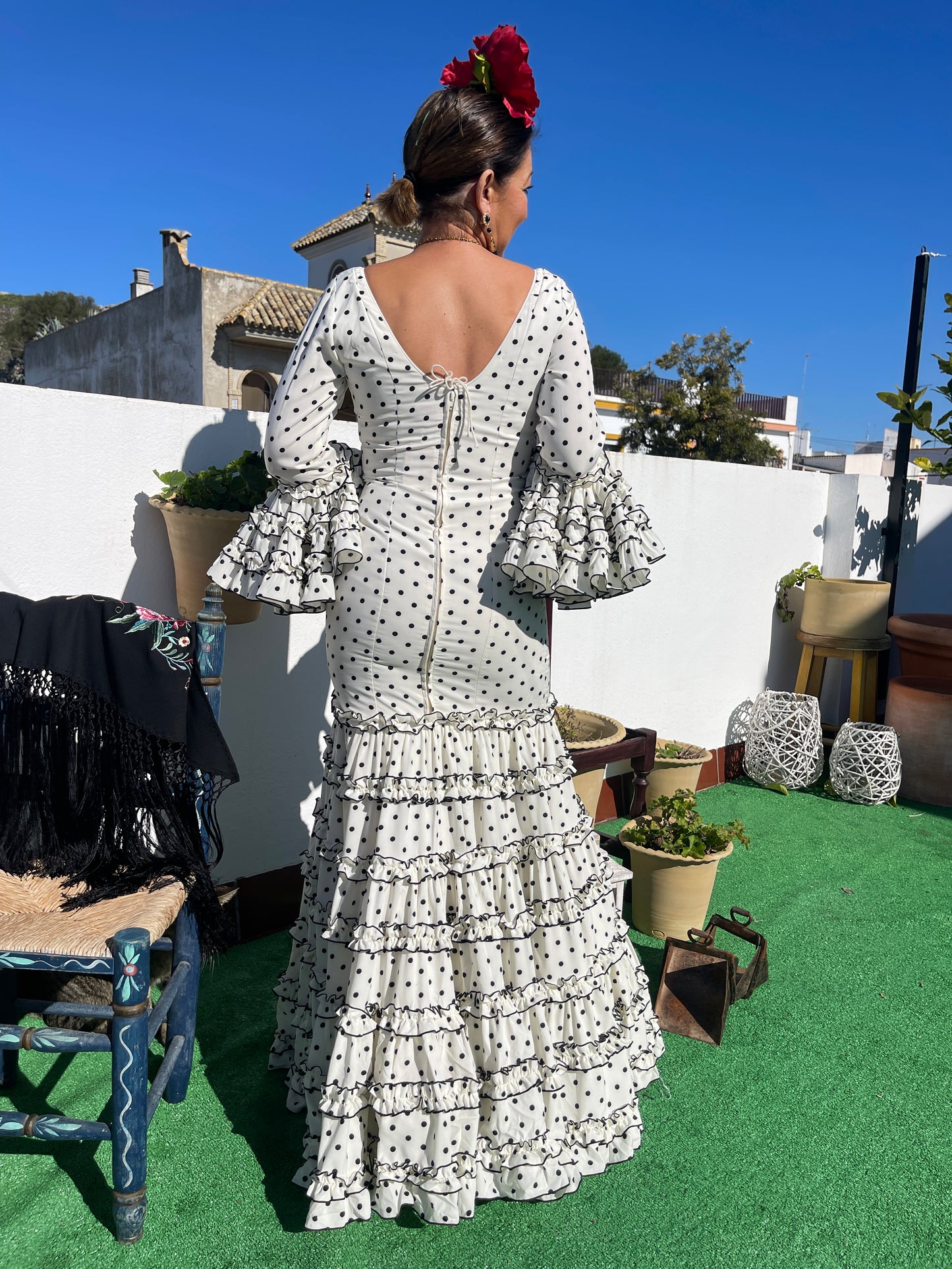 Espalda del vestido de flamenca de mujer con corte canastero blanco con lunar negro. El traje de flamenca de moda en 2024. Ideal para ir guapa y con estilo a la feria de abril, candelarias o el camino del Rocío. De complemento lleva una flor de peonia grande roja y un mantón negro con flecos y flores bordadas apoyado en una silla.