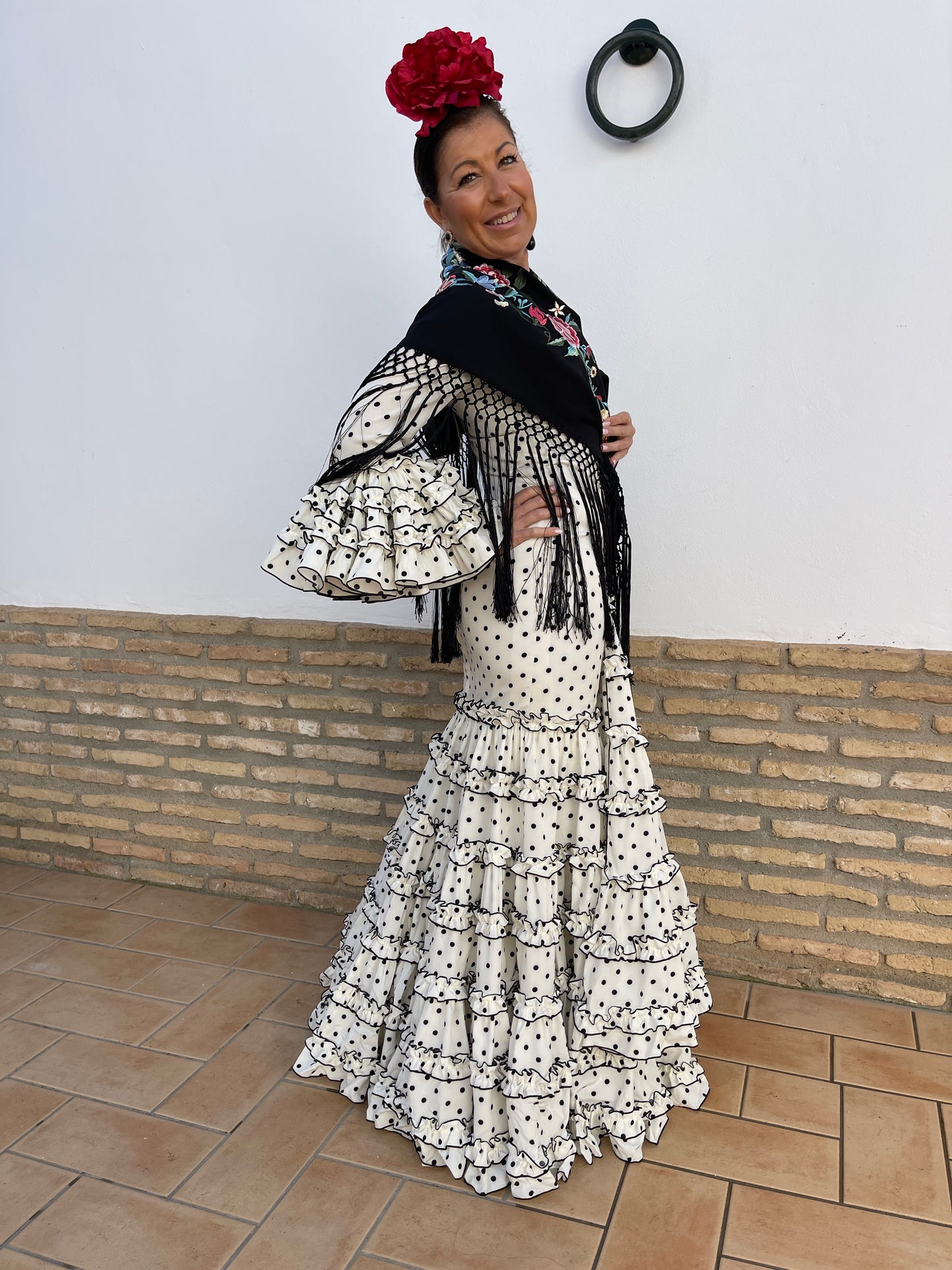 Vestido de flamenca de mujer con corte canastero blanco con lunar negro ideal para la feria de abril, candelarias o el camino de el Rocío. Los complementos son una flor de peonia grande roja, mantón negro con flecos y flores bordadas.