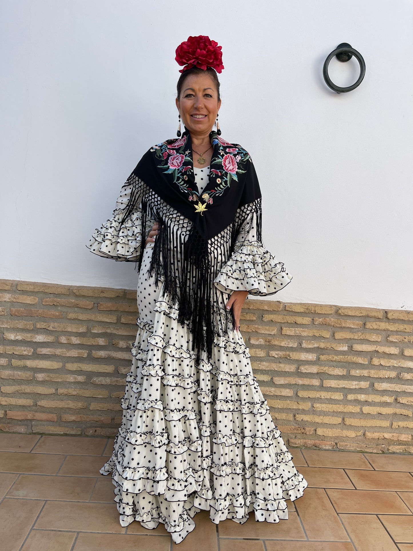 Traje de flamenca de mujer con corte canastero blanco con lunar negro ideal para la feria de abril, candelarias o el camino de el Rocío. Los complementos son una flor de peonia grande roja, pendiente largo dorado y negro en forma de lágrima, mantón negro con flecos y flores bordadas y broche dorado en forma de hoja.