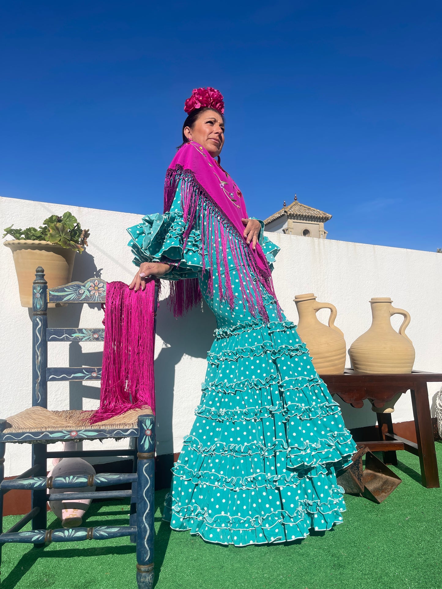 Vestido de gitana turquesa con lunares blancos, mantón bordado flamenca con de flores en color buganvilla y ramillete buganvilla. Este modelo pertenece a la moda flamenca 2024